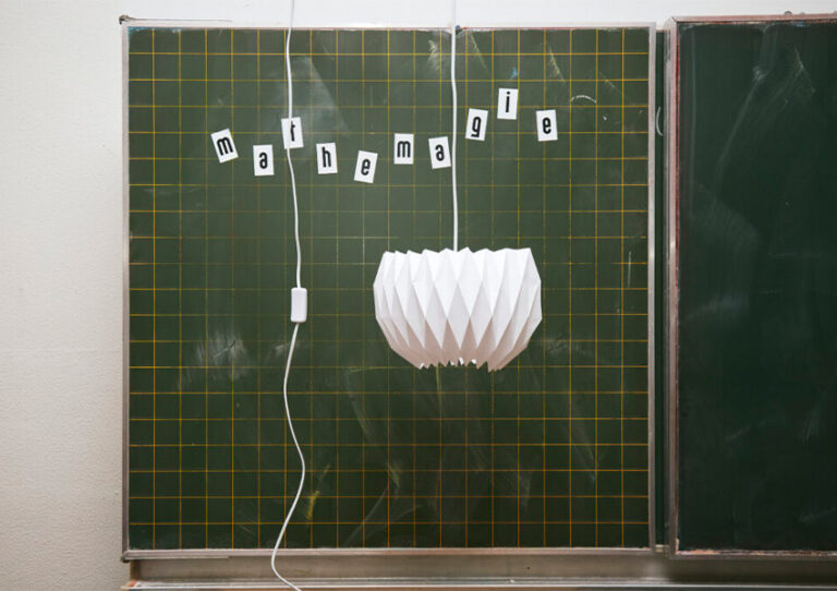 Eine selbstgebaute Lampe hängt vor einer Tafel. Auf der Tafel steht in aufgeklebten Buchstaben: Mathemagie.