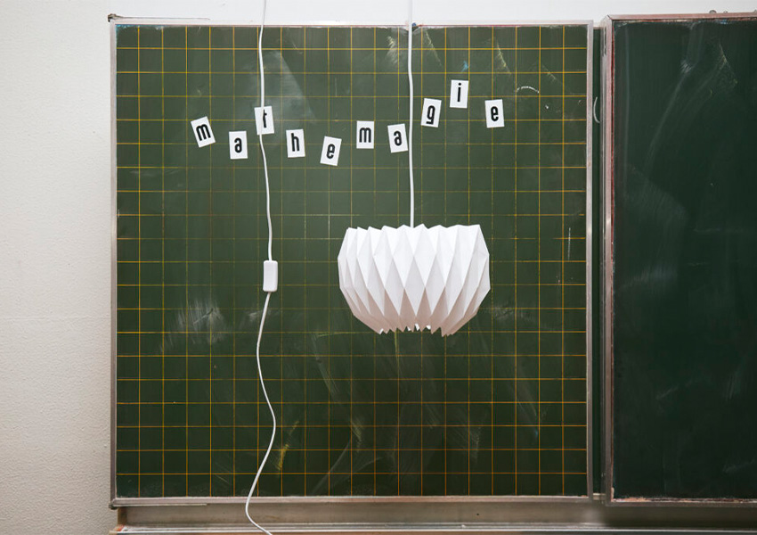 Eine selbstgebaute Lampe hängt vor einer Tafel. Auf der Tafel steht in aufgeklebten Buchstaben: Mathemagie.