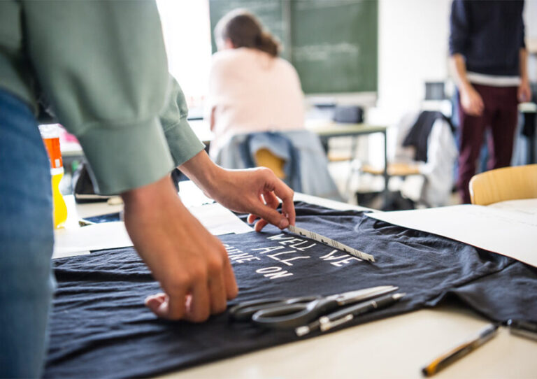 Jemand trägt einen Schriftzug auf ein T-Shirt auf. Daneben liegen Stifte und eine Schere. Auf dem T-Shirt wird stehen: Well we all shine on