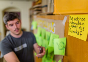 Ein Designer steht an einer Wand mit vielen Post-Its. Vorne hängt eines mit der Frage: Aus welchen Materialien besteht das Produkt?